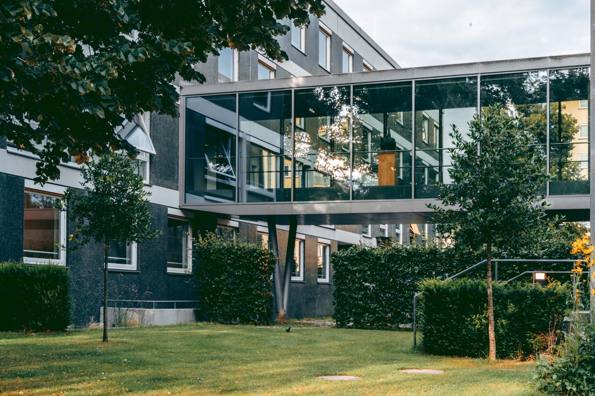 Multi-story building with a glass walkway, a mowed lawn, young trees and clipped dark green hedges
