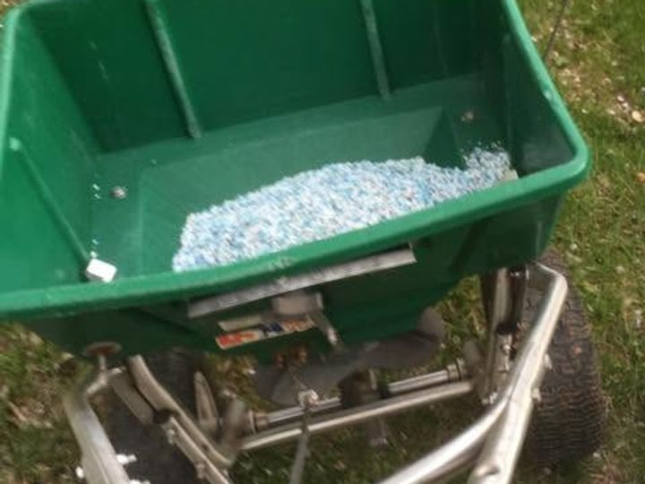 Top view of a green spreader hopper holding blue and white granules beside a blurred wheel