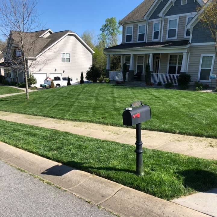 Deep green striped mowed lawn with a black mailbox on a post, a sidewalk and a two-story tan house with a covered porch
