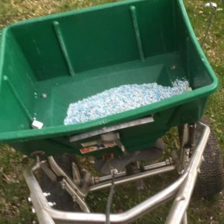 Green walk-behind spreader loaded with blue and white granules on a patchy lawn